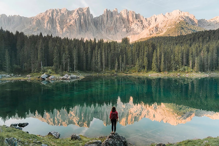 Person standing at the edge of a lake reflecting the surrounding mountains.